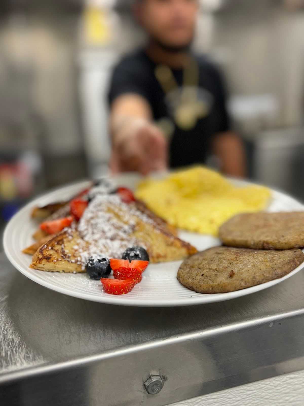 Plated breakfast with French toast, berries, scrambled eggs, and sausage served by a person.