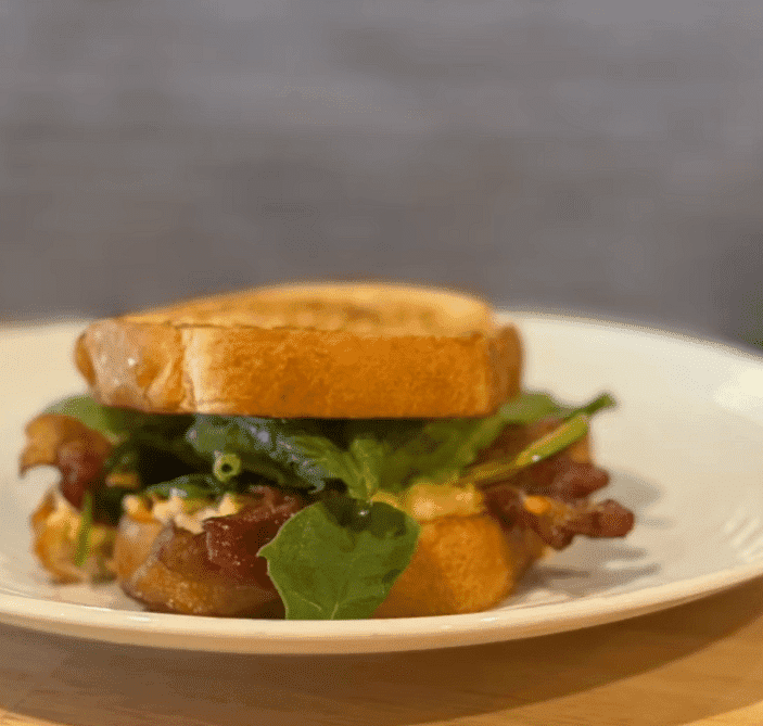 A gourmet sandwich with greens and bacon on a white plate against a blurry background.