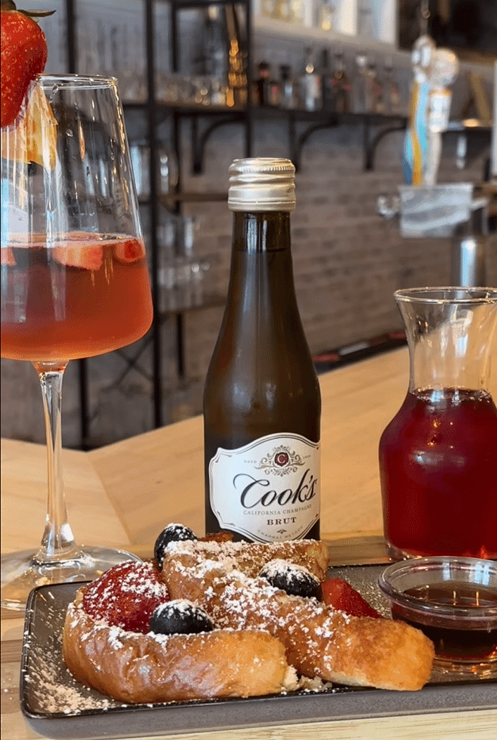 Brunch setting with Cook's Brut, a fruit-topped pastry, a glass of red drink, and decor in the background.