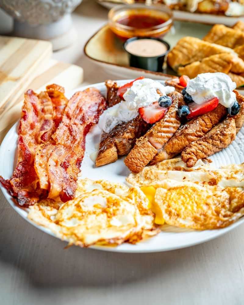 Breakfast plate with bacon, eggs, pancakes, strawberries, and blueberries topped with whipped cream.
