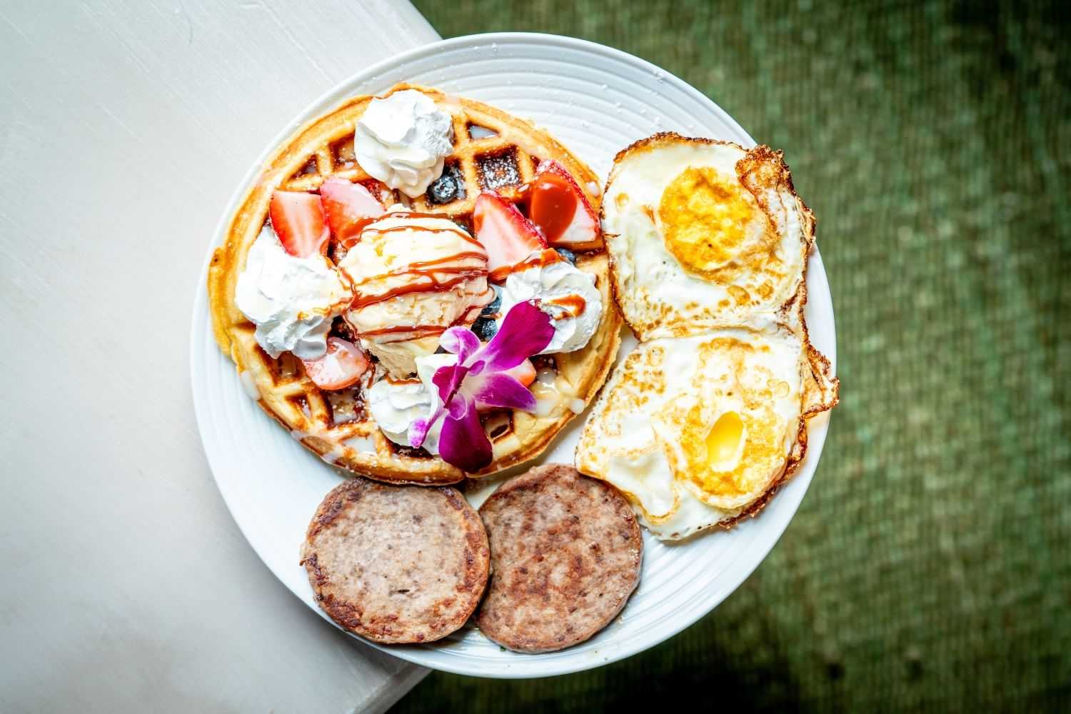 Waffles with strawberries, whipped cream, fried eggs, and sausage on a white plate.