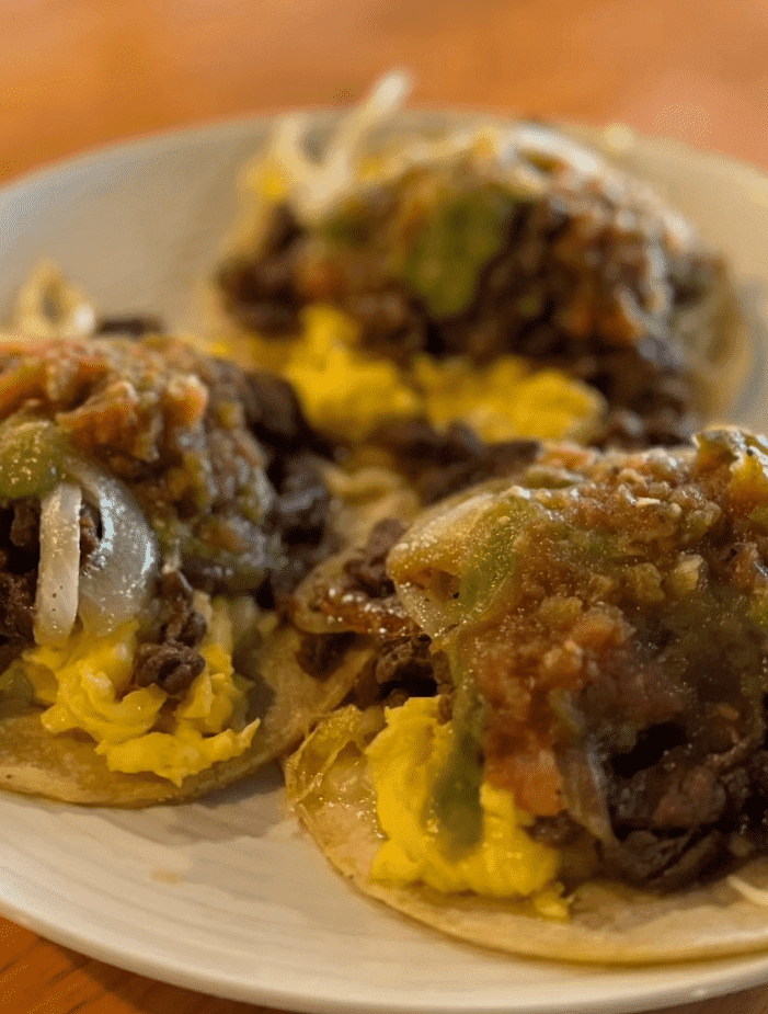 Plate of tacos topped with scrambled eggs, beef, onions, and green and red salsa.