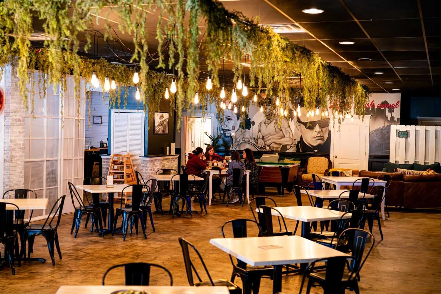 Cozy café with hanging greenery, warm lighting, and patrons enjoying food at tables.